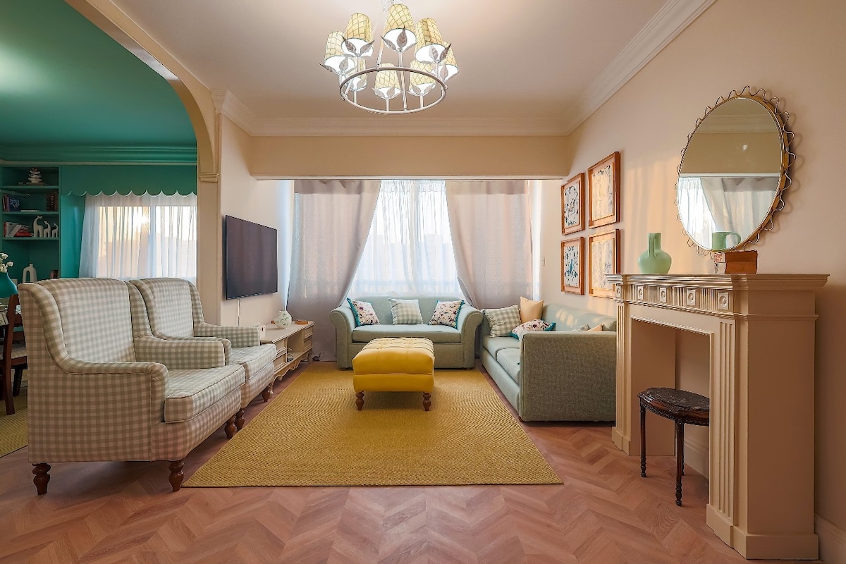 A bright living room features two patterned sofas and a yellow ottoman as a focal point. Soft natural light filters through sheer curtains, illuminating the space. A round mirror and framed artwork add a decorative touch to the walls, while a vintage fireplace adds character.