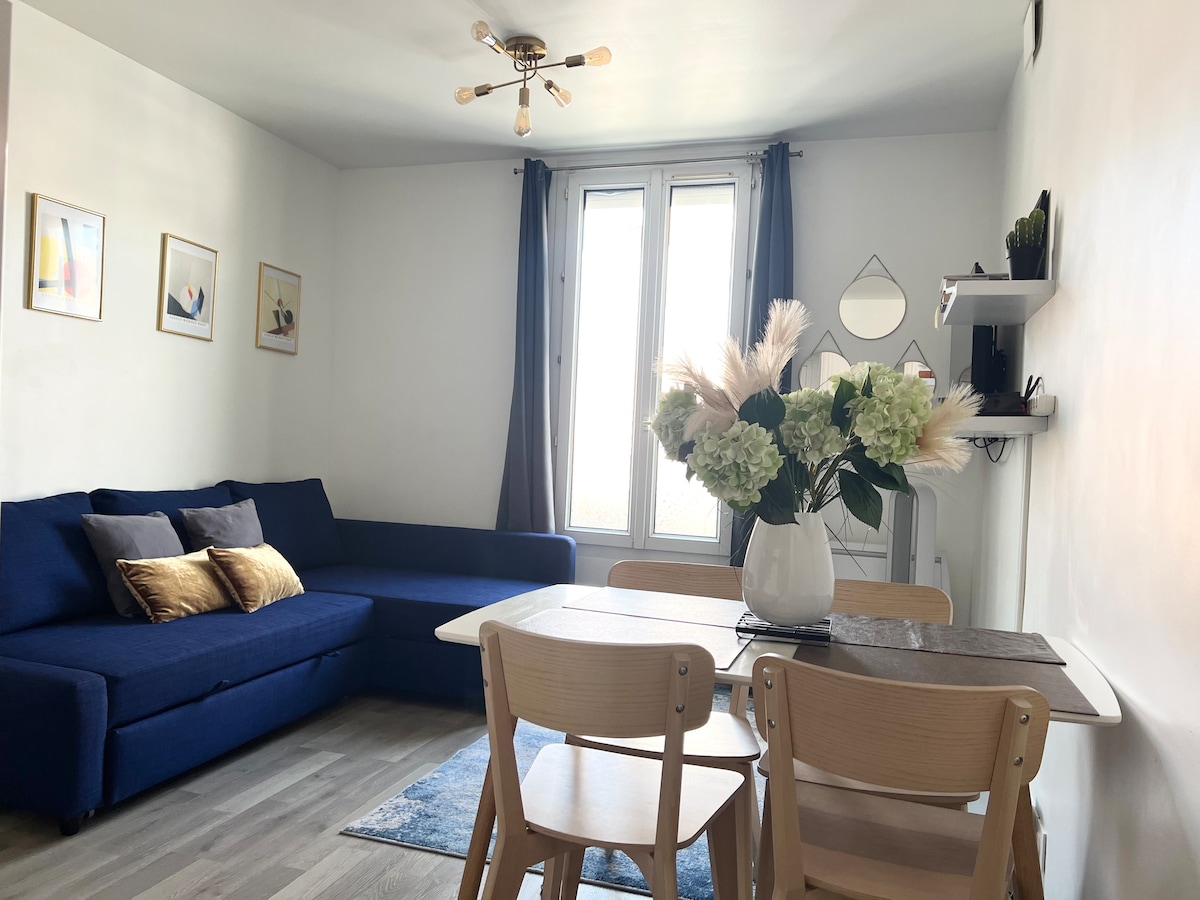 A bright living space is featured, showcasing a dark blue sofa with decorative cushions. A dining table with wooden chairs is positioned beside a window that allows natural light to fill the room. Neutral walls and minimal decor enhance the fresh and modern aesthetic.
