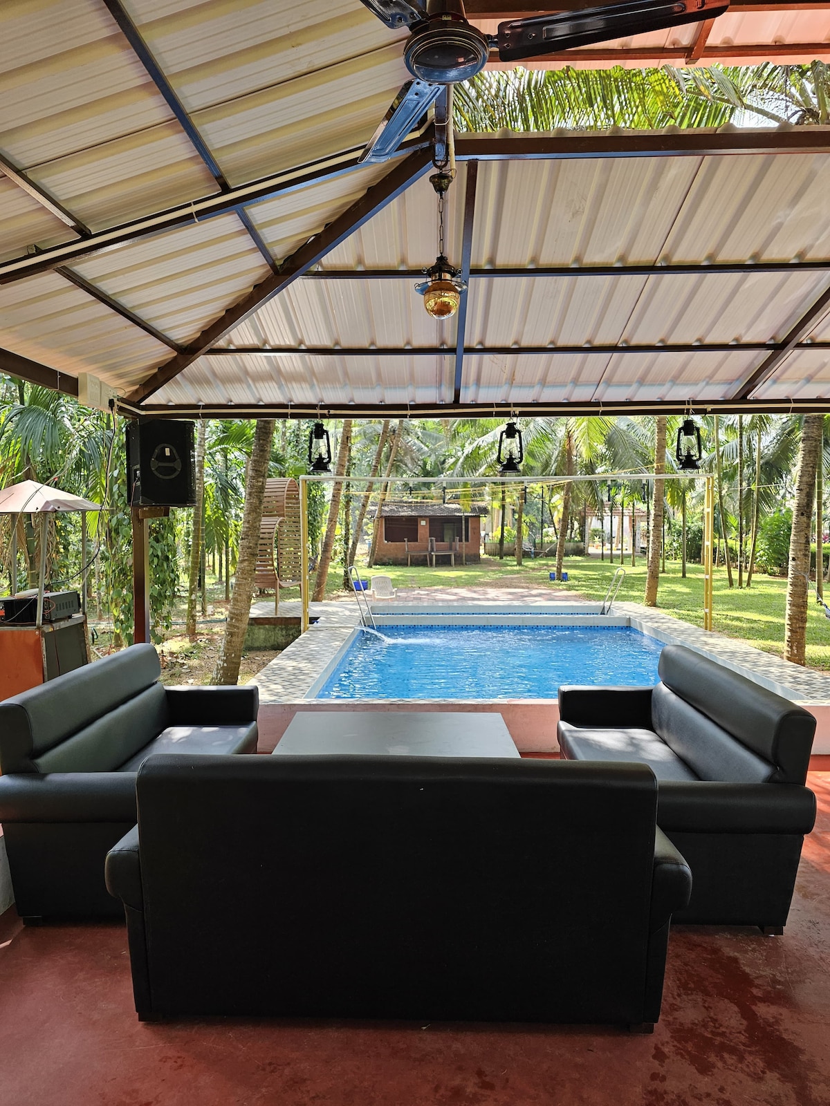 A shaded outdoor seating area features sleek black sofas arranged around a central table. The view overlooks a tranquil pool surrounded by lush palm trees, with additional seating in the background and a charming structure visible beyond the greenery.