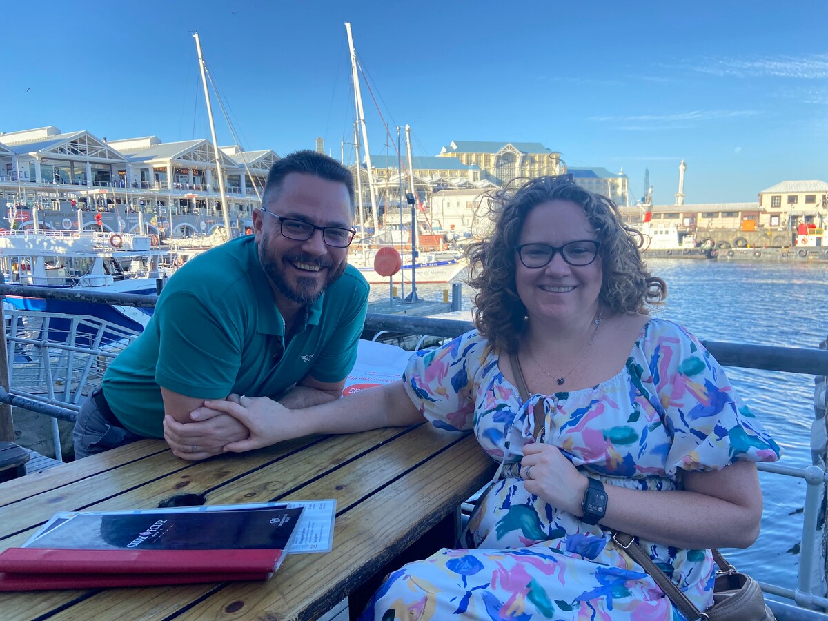 A wooden table is positioned near the water, where two people are seated side by side. Menus and a purse are visible on the table, with boats and the waterfront in the background under a clear blue sky.