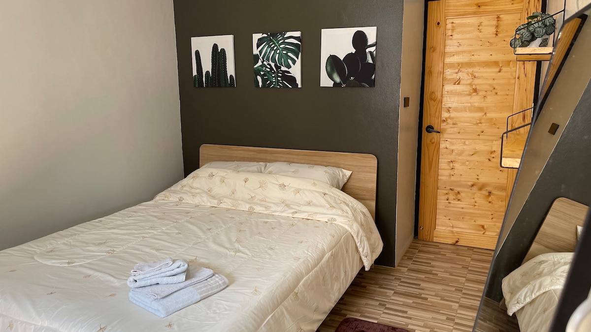 A serene bedroom features a spacious bed with cream-colored linens, flanked by two art pieces depicting plants. Dark green walls create a calming effect, while warm wood accents from the door and staircase add contrast to the overall design.