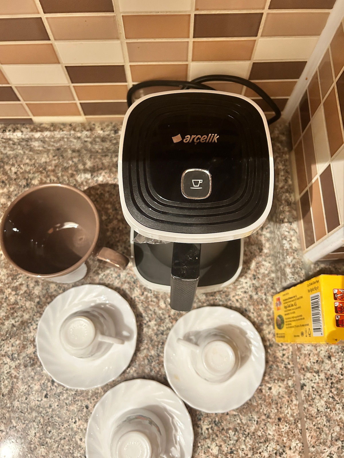 A coffee maker is positioned on a granite countertop, with a single cup beside it. Three white saucers are placed neatly nearby. A brown mug is visible, alongside a packet of coffee. The background features a patterned tile backsplash.