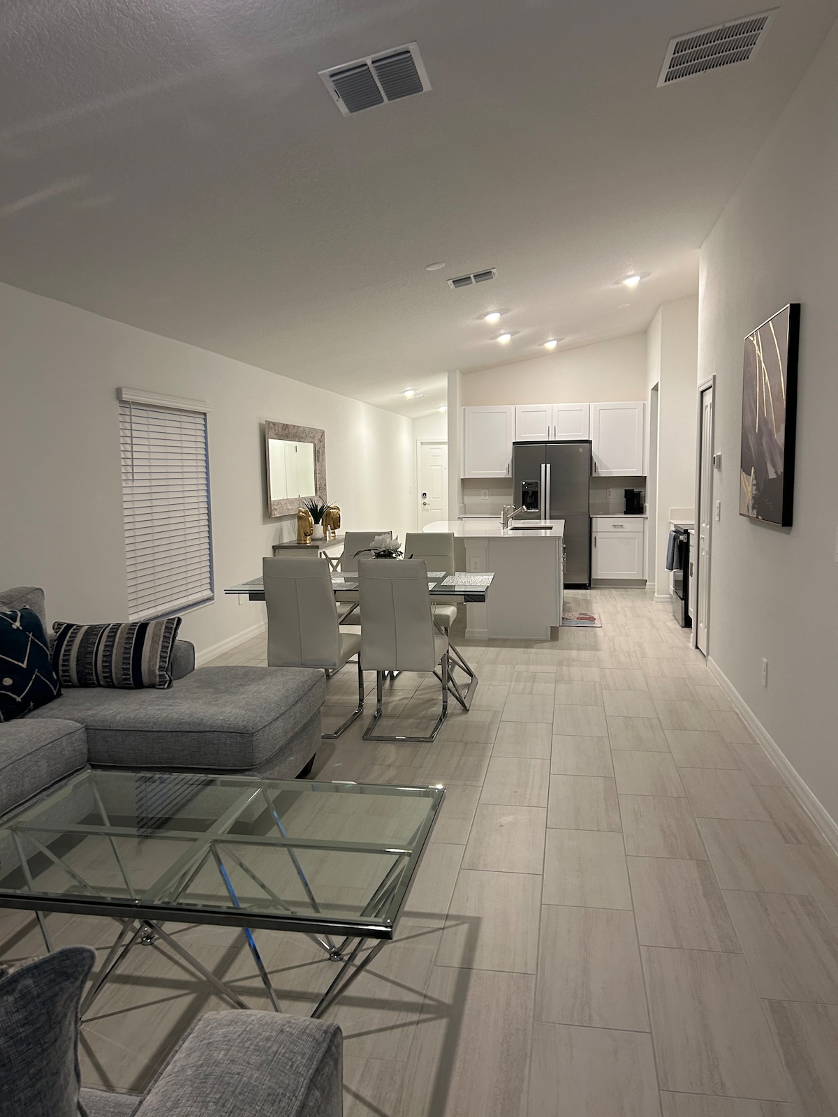 An open living area is showcased, featuring a cozy gray sectional sofa and a glass-top coffee table. A dining table is set with white modern chairs. The kitchen is visible, equipped with white cabinetry and stainless-steel appliances. Natural light filters through the window.