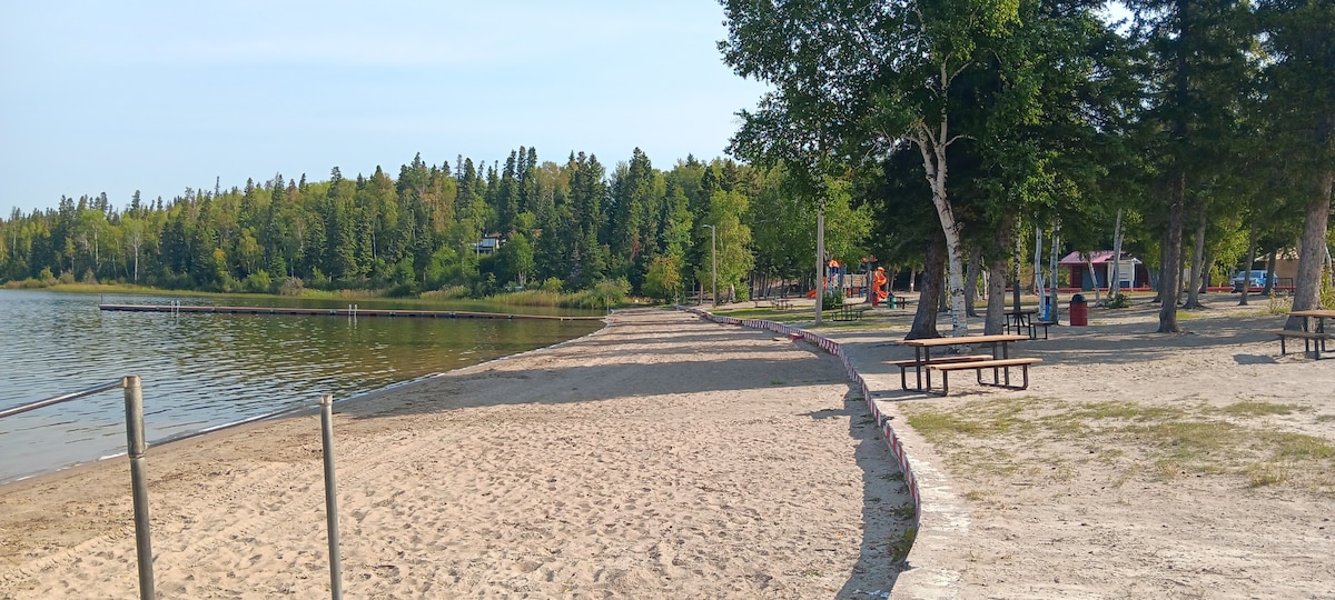 A sandy beach area is shown along the calm water, surrounded by green trees. Picnic tables are placed on the shore, offering space for gatherings. The pathway meanders beside the beach, leading to a playground visible in the background.