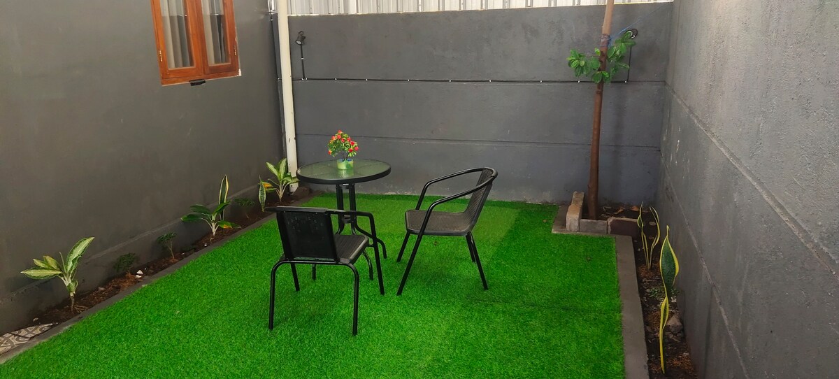 A small garden area features a green artificial lawn bordered by plants. A round table is accompanied by two black chairs. A decorative flower arrangement sits at the center of the table, providing a casual setting for relaxation.