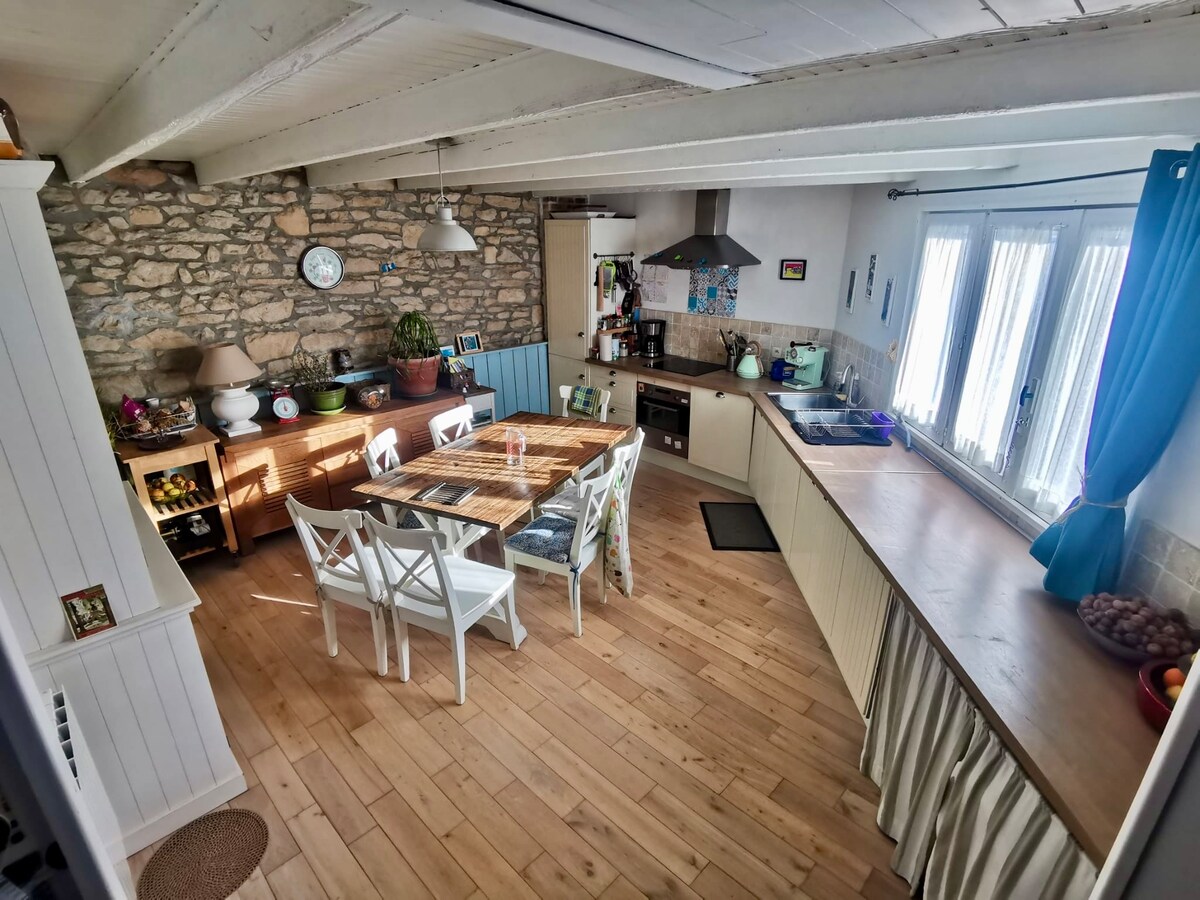 A spacious kitchen and dining area features exposed stone walls and wooden beams. A large dining table with white chairs occupies the center, surrounded by plants and kitchen appliances. Light streams through double windows draped with blue curtains, enhancing the bright and airy feel of the space.