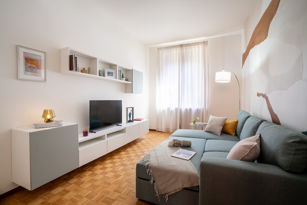 A bright living area features a large, comfortable sectional sofa adorned with soft throw pillows. A modern TV stand holds a flat-screen television against a light-colored wall. Natural light filters in through sheer curtains, illuminating a cozy reading nook with decorative shelves.