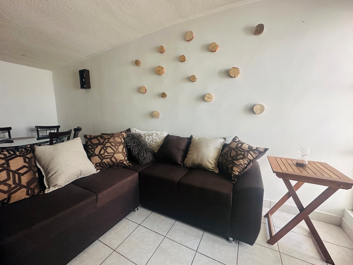 A cozy living area features a dark sectional sofa adorned with various patterned cushions. Natural elements are incorporated into the decor through decorative wooden pieces mounted on the wall. A small wooden side table complements the seating arrangement, creating a comfortable space for relaxation.