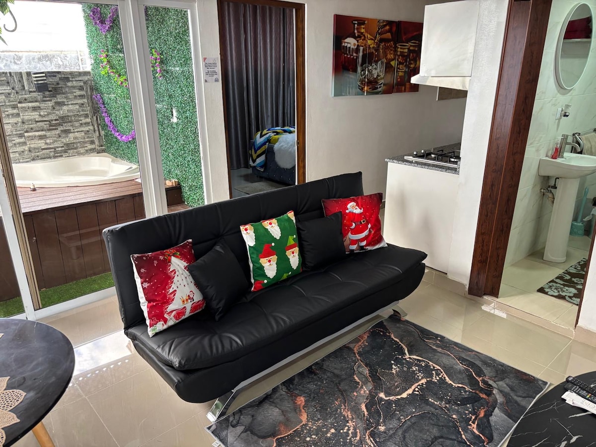 A modern living area features a sleek black sofa adorned with festive pillows. Nearby, large glass doors open to an outdoor space with greenery and a private jacuzzi. A stylish rug complements the room's decor, while a kitchen area is visible in the background.