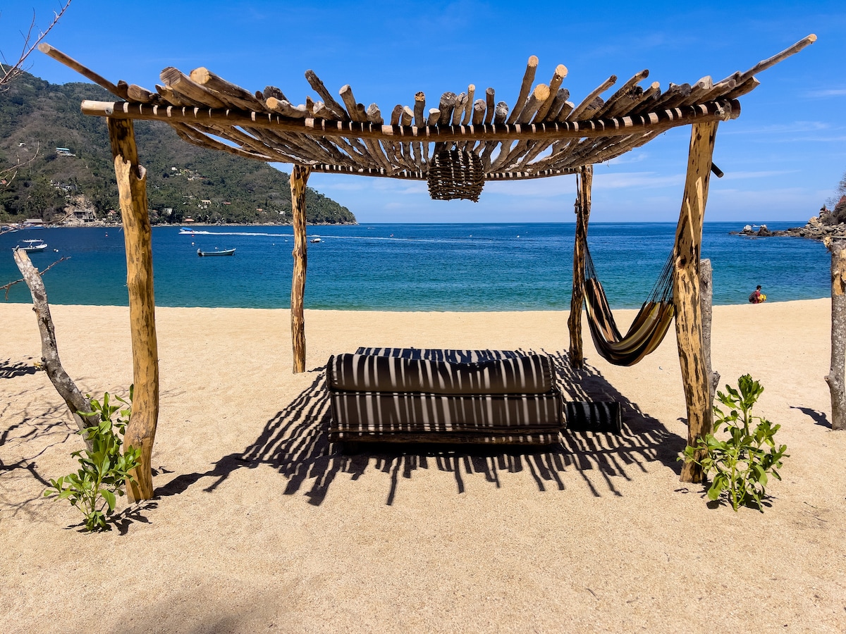 A shaded beachfront structure crafted from natural materials features a striped seating area and a hammock. Soft sand stretches in front, leading to calm, blue waters. Rows of shadow create a serene ambiance, with distant boats visible on the horizon.