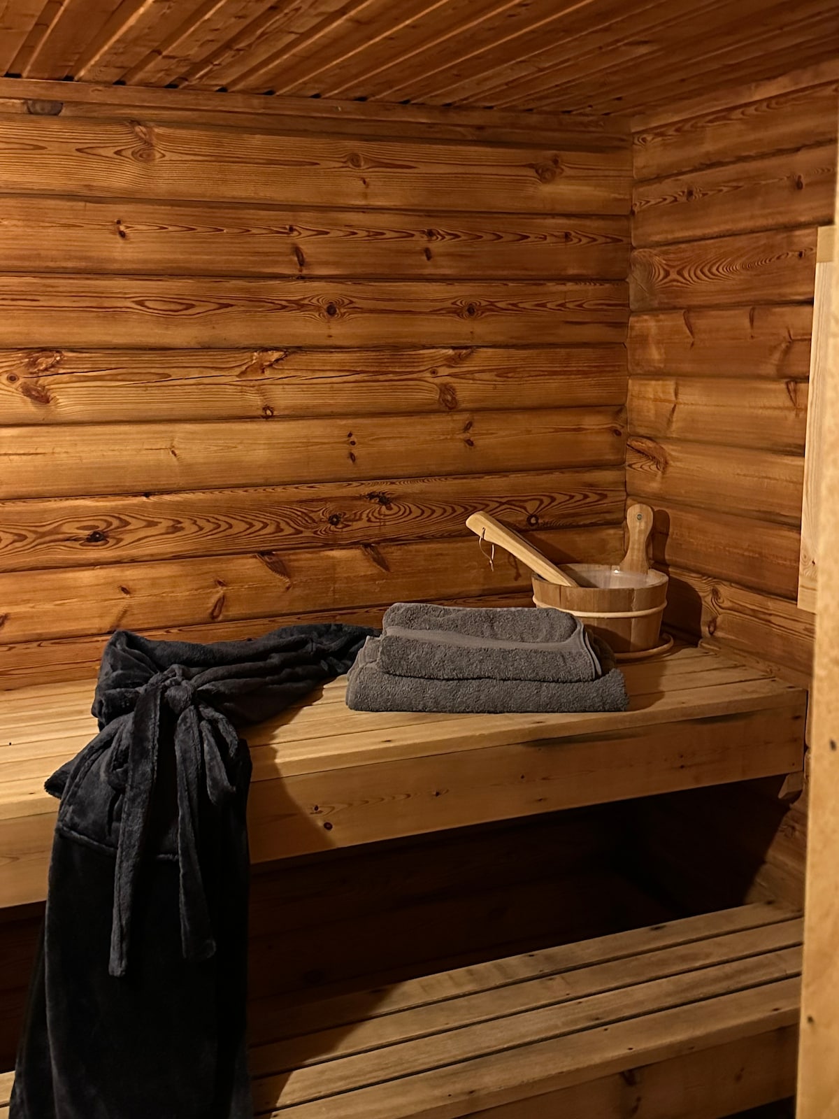 The sauna interior features warm wooden panels and a built-in wooden bench. Soft gray towels are neatly arranged, alongside a wooden bucket and ladle. A dark robe is draped over the bench, inviting relaxation in a serene and cozy environment.