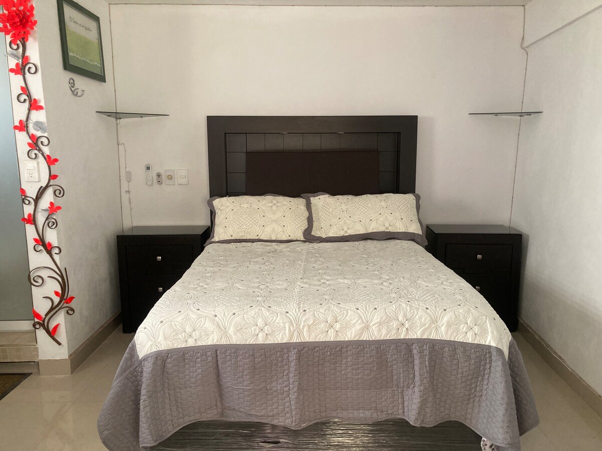 A spacious room features a large bed with an intricately designed quilt. Dark wood nightstands are positioned on either side, providing additional storage. Soft lighting accentuates the neutral color scheme, while the tiled floor adds a clean, modern touch to the space.