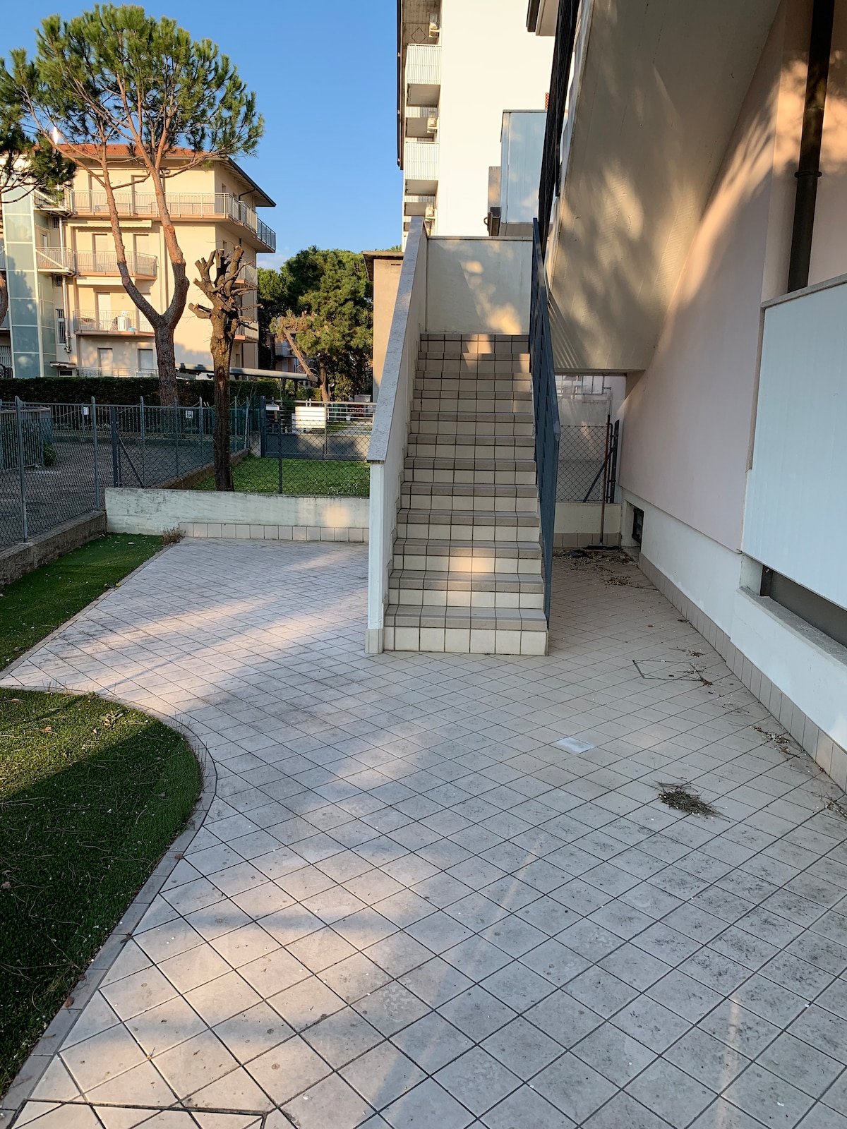 A pathway leads to a staircase, providing access to the first floor of the building. The area is bordered by greenery, with a clear view of adjacent structures and a fenced section in the background.