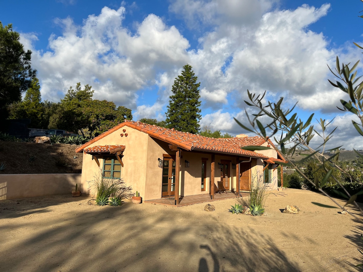 The casita features a charming Spanish style exterior with a tiled roof and smooth stucco walls. Custom wooden doors open onto a patio surrounded by native landscaping. The peaceful surroundings include views of the hills and scattered vegetation, emphasizing the serene atmosphere.