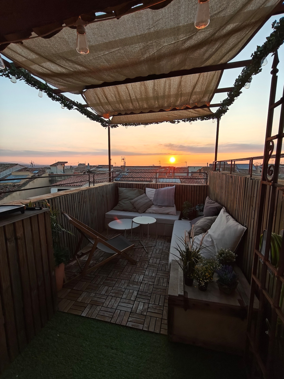 A panoramic terrace features comfortable seating and stylish decor, surrounded by wooden panels and plants. The sunset creates a warm glow over the rooftops of Aix-en-Provence, providing a serene backdrop for relaxation.