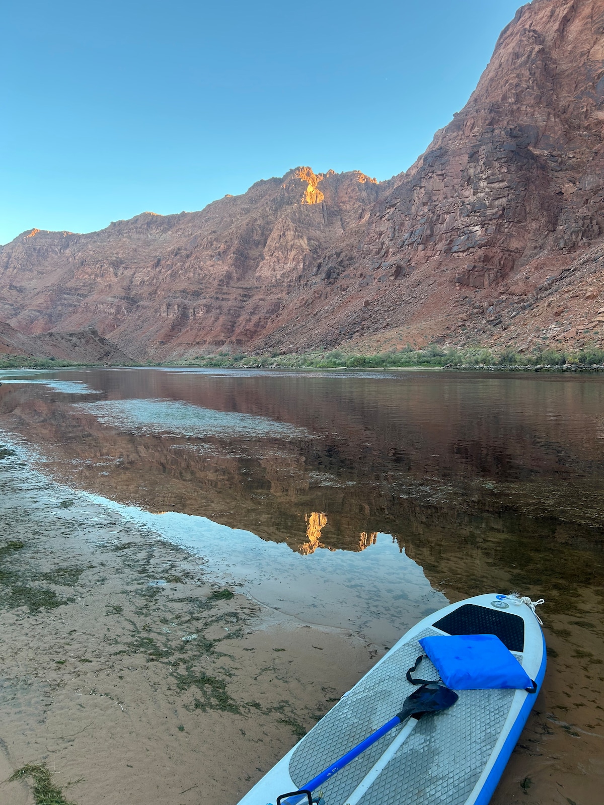 A serene riverside scene is captured, with towering red rock cliffs reflected in the calm water. A paddleboard rests on the sandy shore, while lush greenery peeks from the base of the cliffs. The sky above is clear and bright, enhancing the peaceful setting.