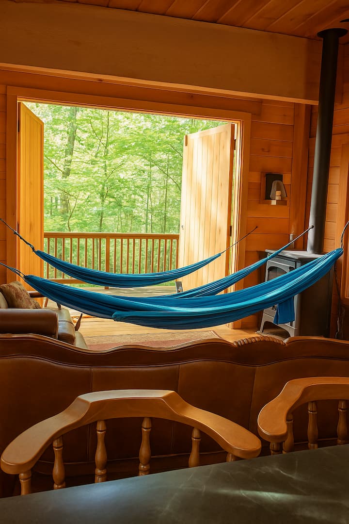 Tre amache blu possono essere appese a calde pareti di legno e una stufa a legna nera. Le grandi porte doppie si aprono su una terrazza che si affaccia su alberi verdi e rigogliosi, riempiendo lo spazio di luce naturale e offrendo una vista tranquilla sulla foresta.