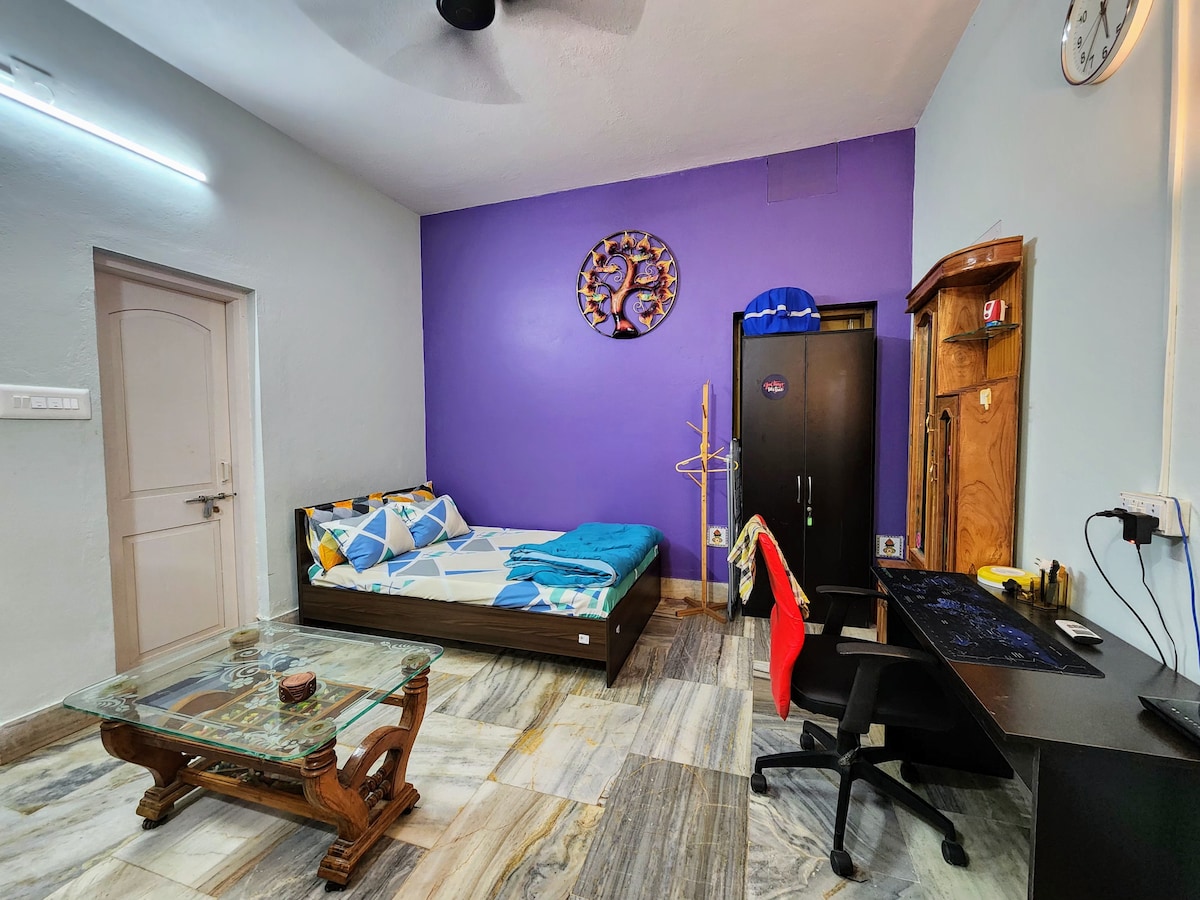 A private studio room features a spacious bed with a colorful blanket, set against a rich purple accent wall. A glass coffee table is positioned nearby, alongside a wooden desk equipped with a computer and a black chair. Wardrobe and wall decorations add to the room's functionality.