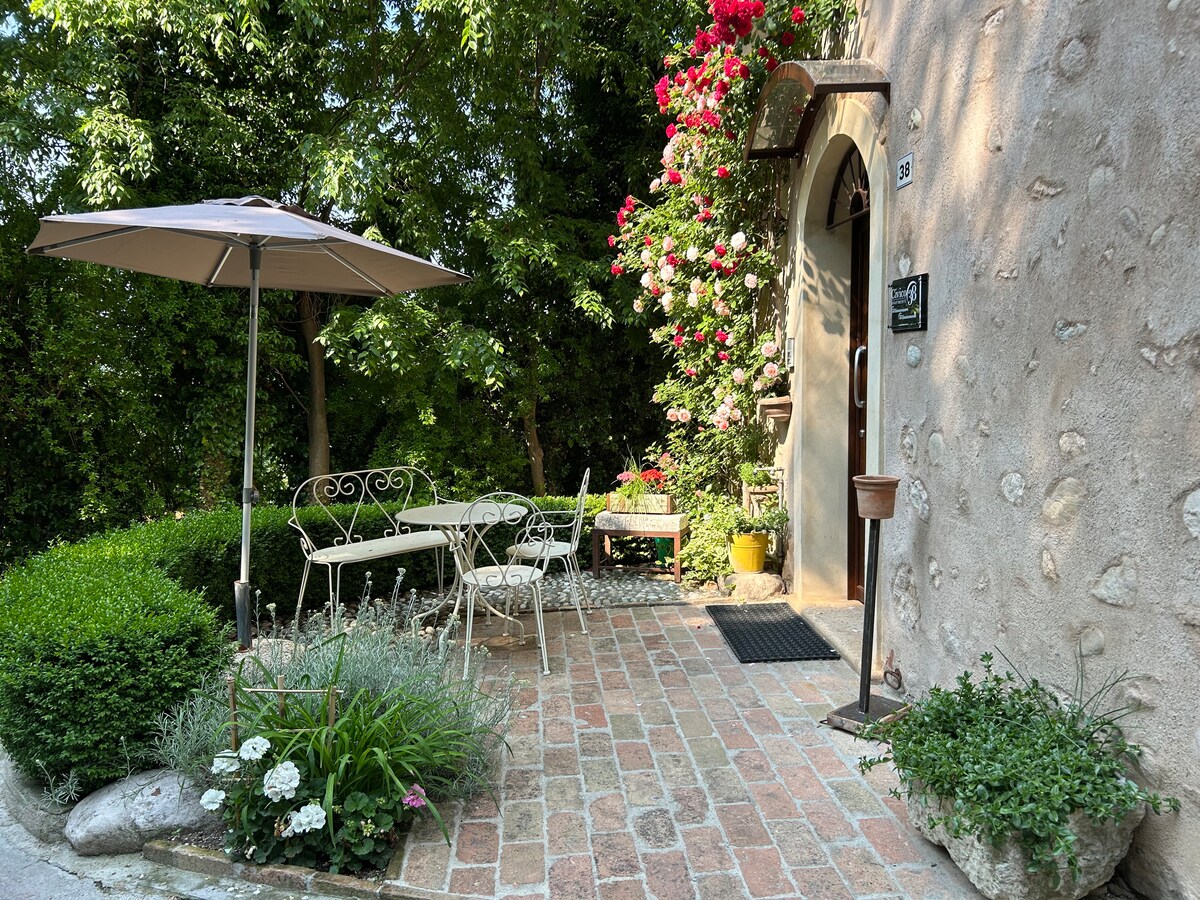 A charming exterior area is depicted, featuring a small table and two chairs under an umbrella. Vibrant flowers climb the wall, and well-tended greenery accents the brick pathway, creating a serene setting. A welcoming entrance is visible, set against a rustic stone wall.