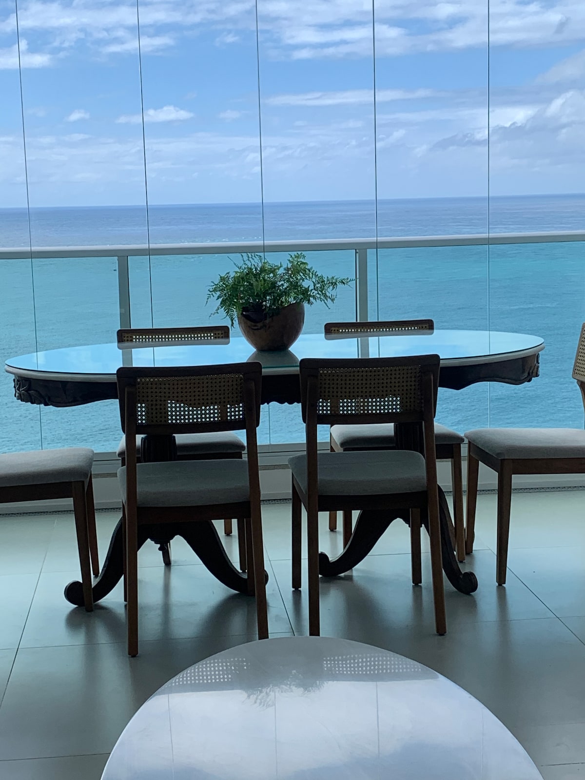 A dining table with a glass top is set against a backdrop of panoramic ocean views. Four wooden chairs surround the table, and a potted plant adds a touch of greenery. Large windows allow natural light to illuminate the space.
