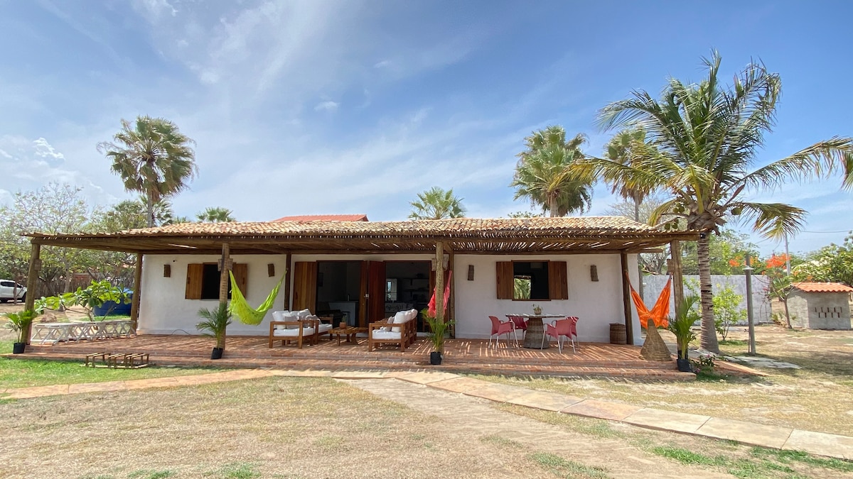 An inviting exterior view of a spacious chalet is presented, showcasing a covered terrace adorned with colorful hammocks. Comfortable seating areas are arranged under a thatched roof, framed by palm trees and a lush garden, which enhances the outdoor living space.