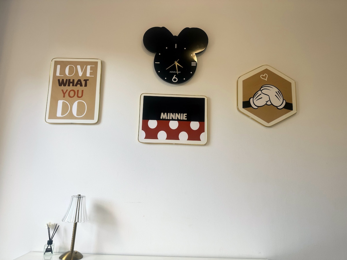 A light-colored wall features three framed decorative pieces. A wall clock in the shape of a mouse head is positioned above a sign with 'MINNIE' and a red polka dot design. A motivational quote 'LOVE WHAT YOU DO' is also displayed, accompanied by a simple table lamp.