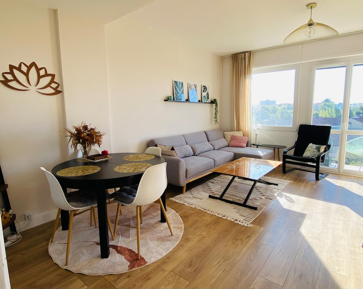 A spacious living area features a comfortable sofa, a round dining table set for four, and a coffee table positioned on a decorative rug. Large windows offer abundant natural light and a view of the surrounding landscape.