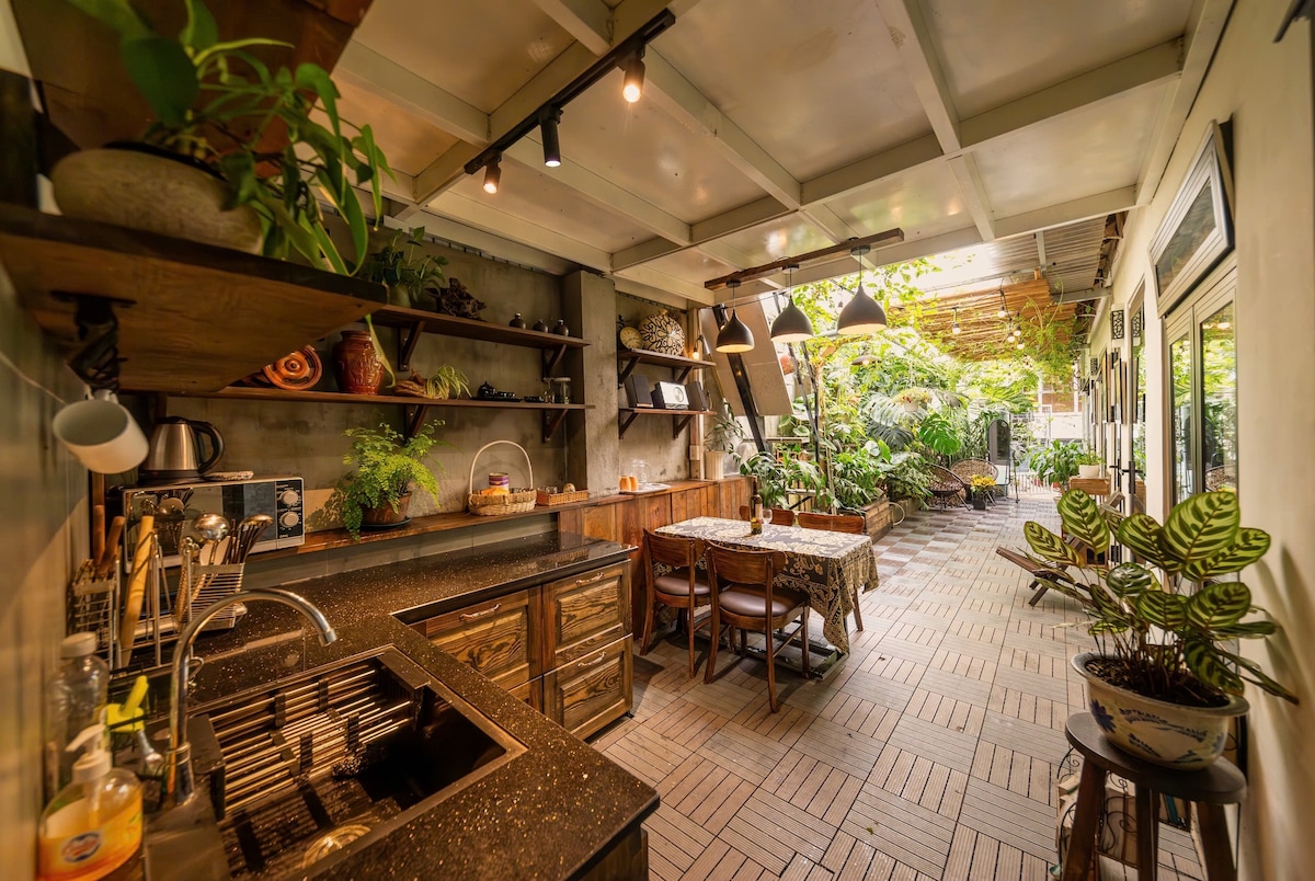 An outdoor kitchen area is surrounded by lush greenery, featuring wooden shelving adorned with plants and various kitchen items. A long table with patterned cloth is positioned for dining, illuminated by overhead lighting. The space offers a calm and inviting atmosphere for meal preparation and gatherings.