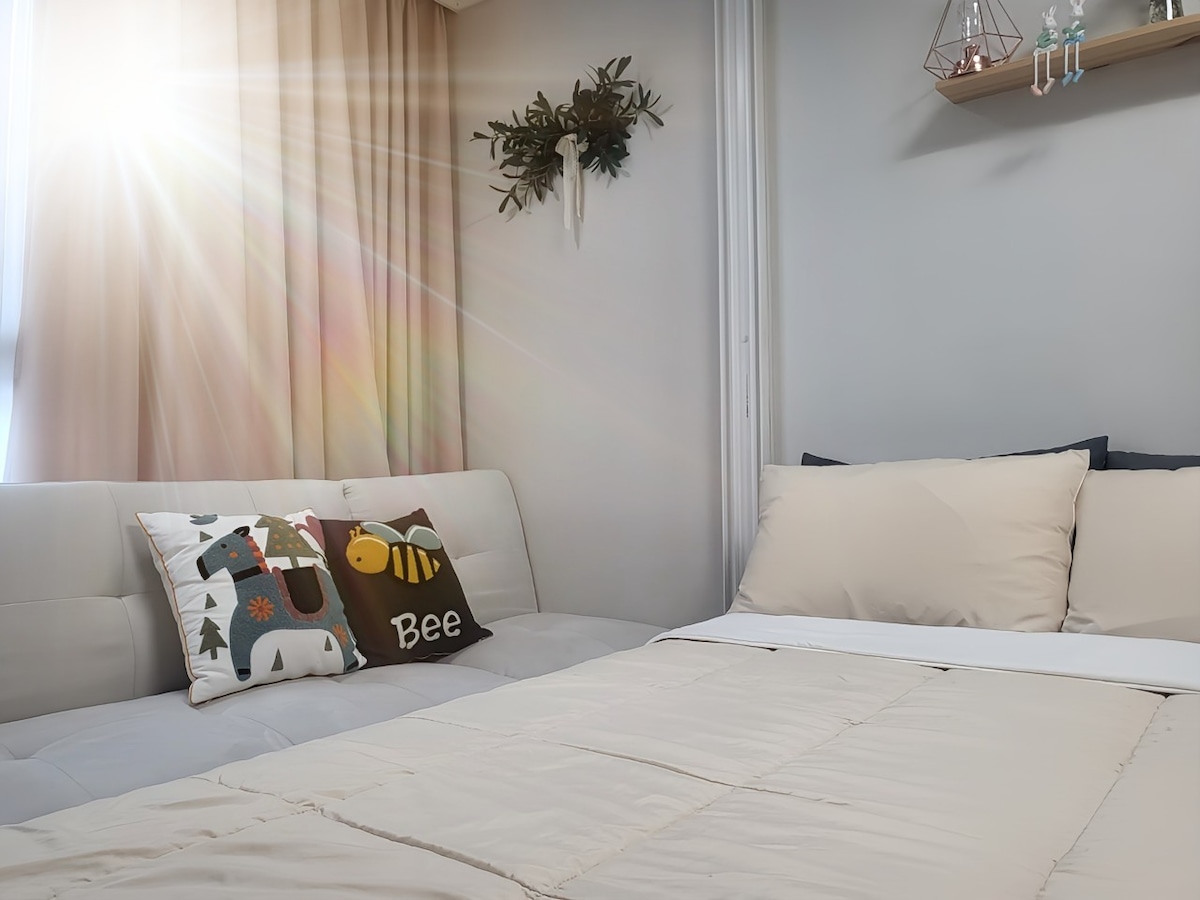 A cozy sleeping area features a spacious bed dressed in soft neutral bedding. A light-colored sofa with decorative cushions adds comfort. Natural light filters in through the sheer curtains, creating a warm atmosphere, while a wall shelf displays decorative items.