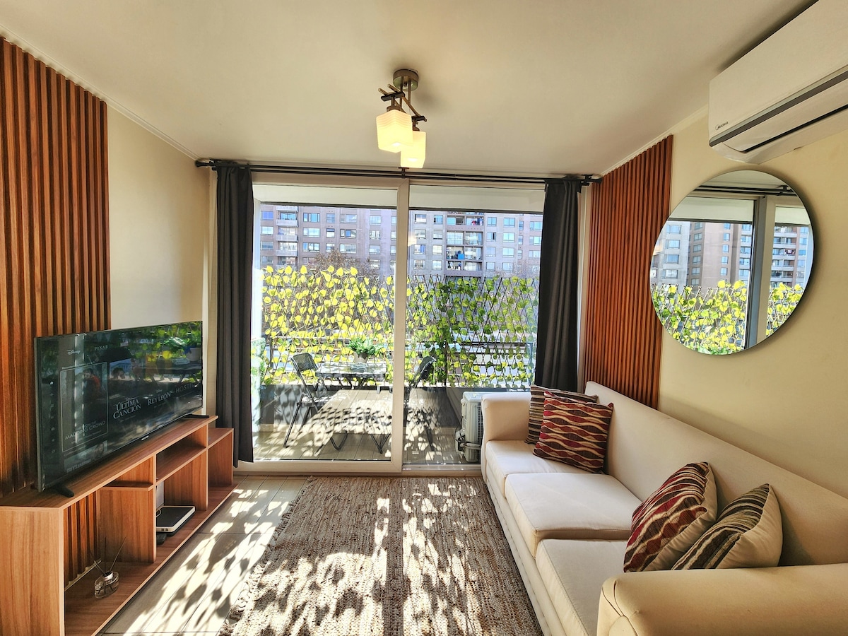 A bright living area is equipped with a comfortable sofa and a modern smart TV. Large sliding glass doors provide access to a balcony, while a round mirror enhances the visual space. Natural light fills the room, highlighting the wood paneling and decor.