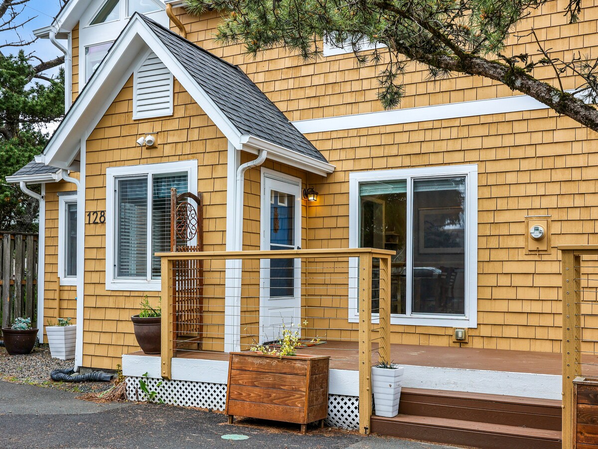 The exterior of a charming coastal house features warm yellow shingles and a welcoming front porch. Fresh plants are arranged in pots, complemented by a wooden planter box. Large windows allow bright natural light, offering a glimpse of the inviting interior.