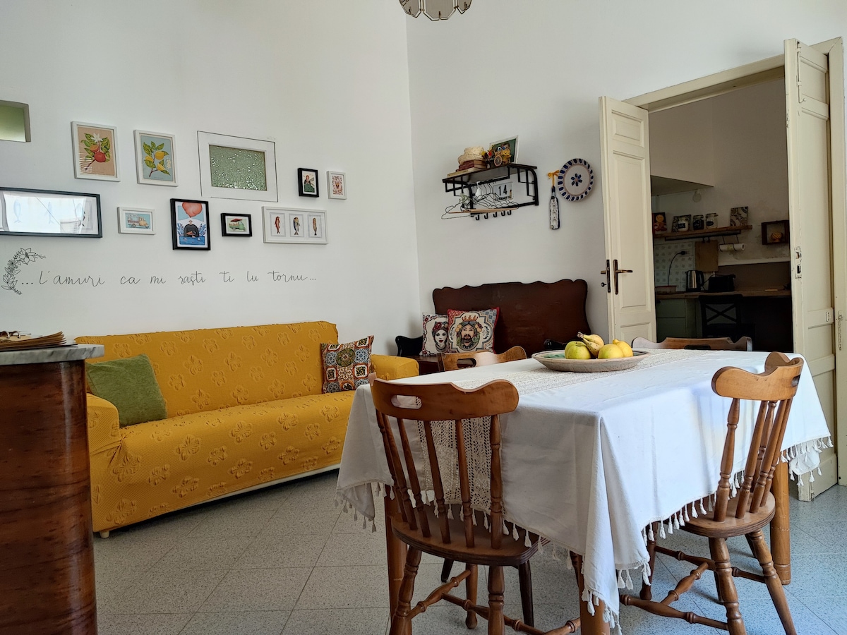 A bright living area features a yellow sofa adorned with colorful pillows. A dining table is set with a white tablecloth, surrounded by wooden chairs. Walls display various framed artworks, adding a personal touch. The open doorway leads into a kitchen space.