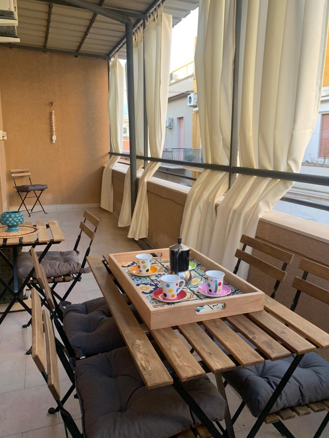 A shaded terrace is furnished with wooden tables and chairs, creating a space for dining or relaxation. A tray holds an assortment of colorful cups and a bottle, suggesting a setting for enjoying beverages. Light filters through flowing curtains, adding a serene ambiance.