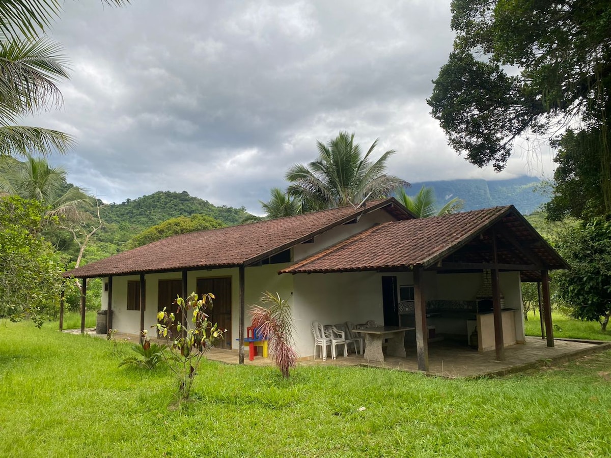 A single-story house is nestled in a green landscape, surrounded by trees and mountains. The structure features a sloped, tiled roof and a covered patio area with outdoor seating. A spacious lawn enhances the peaceful ambiance of the setting.