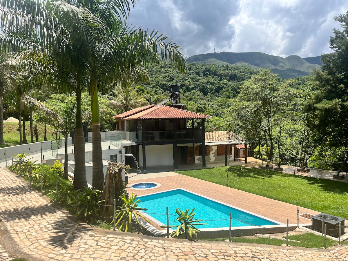 The exterior of a spacious house is surrounded by lush greenery, showcasing a vibrant pool in the foreground. A well-maintained lawn and palm trees frame the scene, while mountains rise in the background beneath a partly cloudy sky.