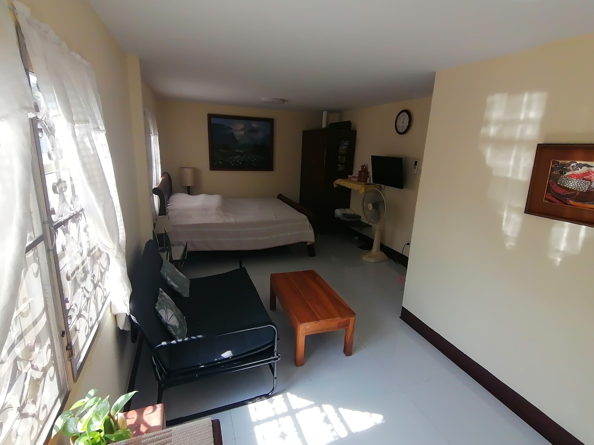 A cozy sleeping area features a large bed alongside a simple wooden coffee table and a seating arrangement. Natural light filters through sheer curtains, illuminating the light-colored walls. An artwork hangs on one wall, adding a touch of character to the space.