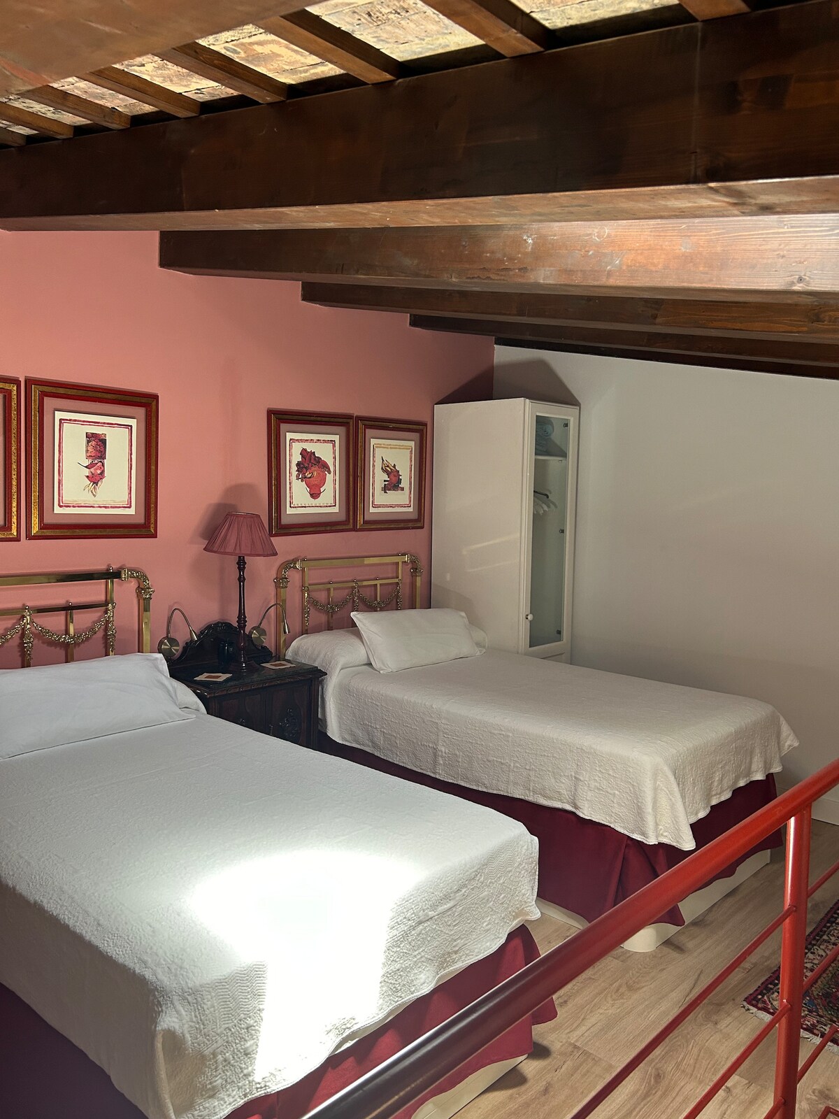 A spacious suite features two separate beds with crisp white linens and decorative bed skirts. Artwork adorns the pink wall, complemented by wooden beams on the ceiling. A wardrobe is visible, providing additional storage space.