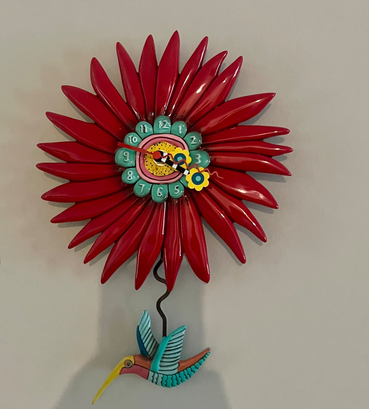 A colorful clock is mounted on the wall, designed as a flower with petal details in vibrant red. The clock features a round face with numbers in contrasting colors and a small bird figure at the bottom, adding a whimsical touch to the decor.