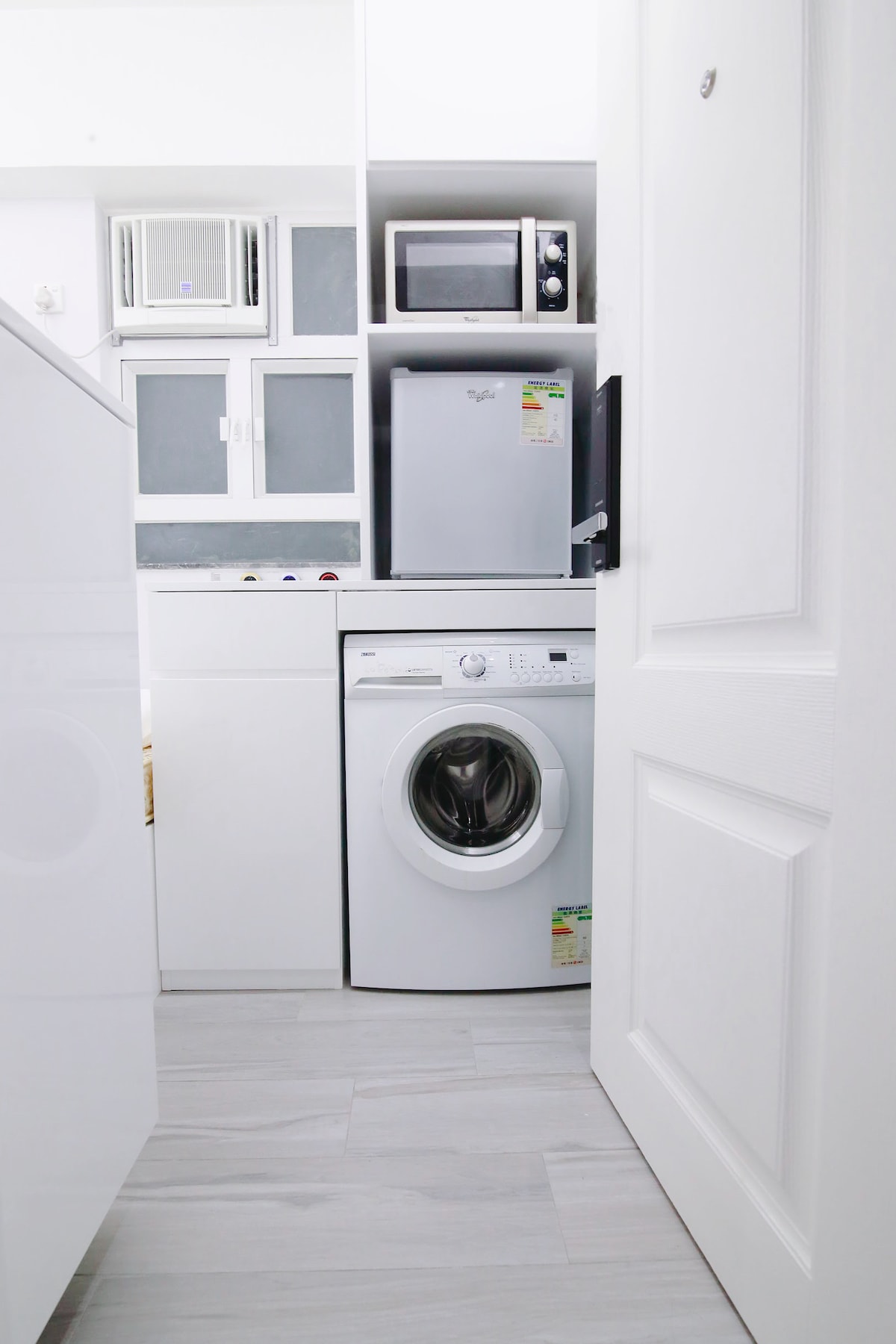 A compact laundry area features a washing machine positioned beneath a countertop. Above the washer, a microwave and a small refrigerator are displayed within built-in shelving. The space is designed with white cabinetry and light flooring, creating a bright, functional atmosphere.
