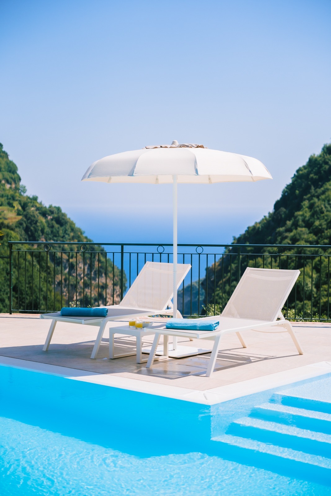 A sunlit pool area is framed by lush green hills and a tranquil ocean backdrop. Two lounge chairs are positioned near the water, accompanied by a large, white umbrella, creating a relaxing space for guests.