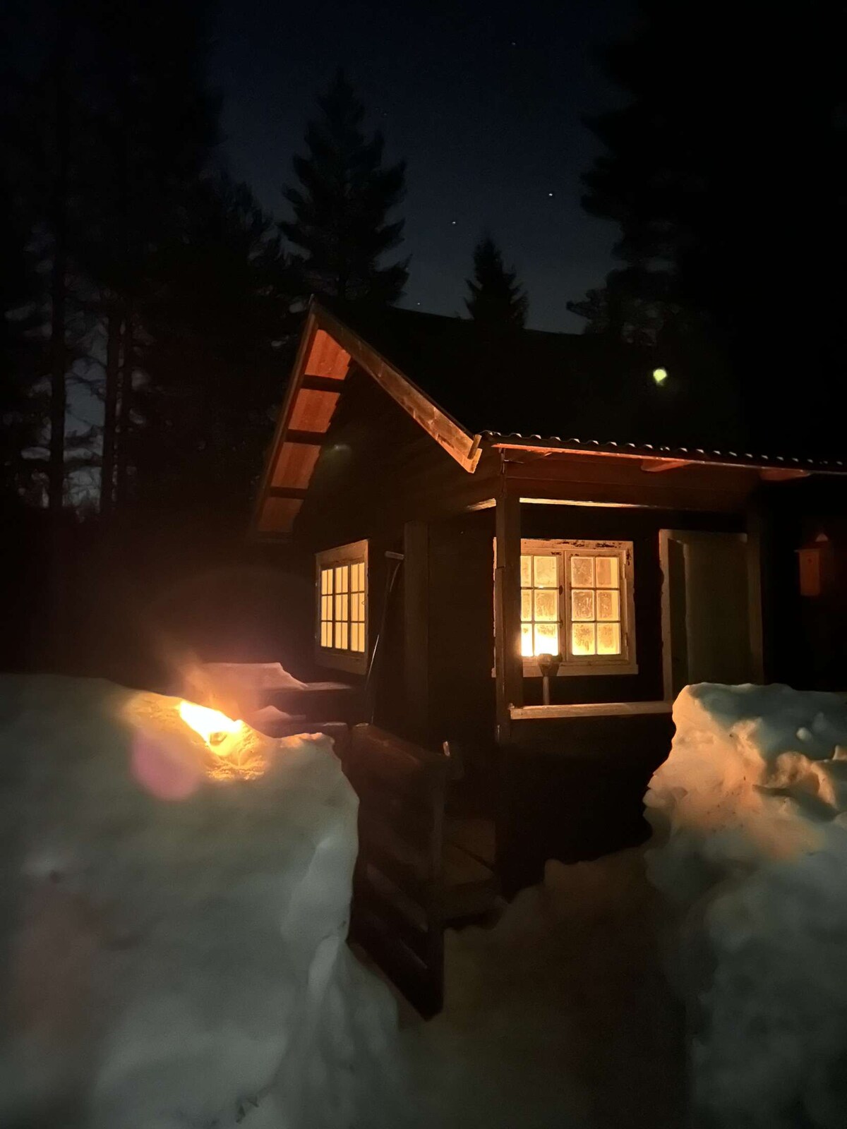 A cabin is illuminated by warm light from windows, surrounded by a snowy landscape. The entrance is visible, featuring steps leading up to the door. Dark trees frame the scene against a night sky dotted with stars.