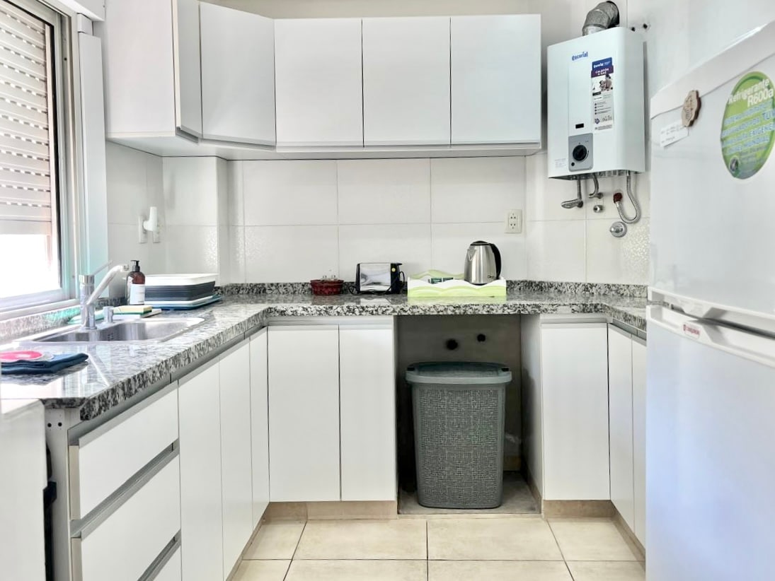 A modern kitchen features white cabinetry and a granite countertop, with essential appliances including a kettle and refrigerator. A light-colored tiled floor complements the space, and natural light enters through the window, illuminating the area and providing a clean and functional setting.