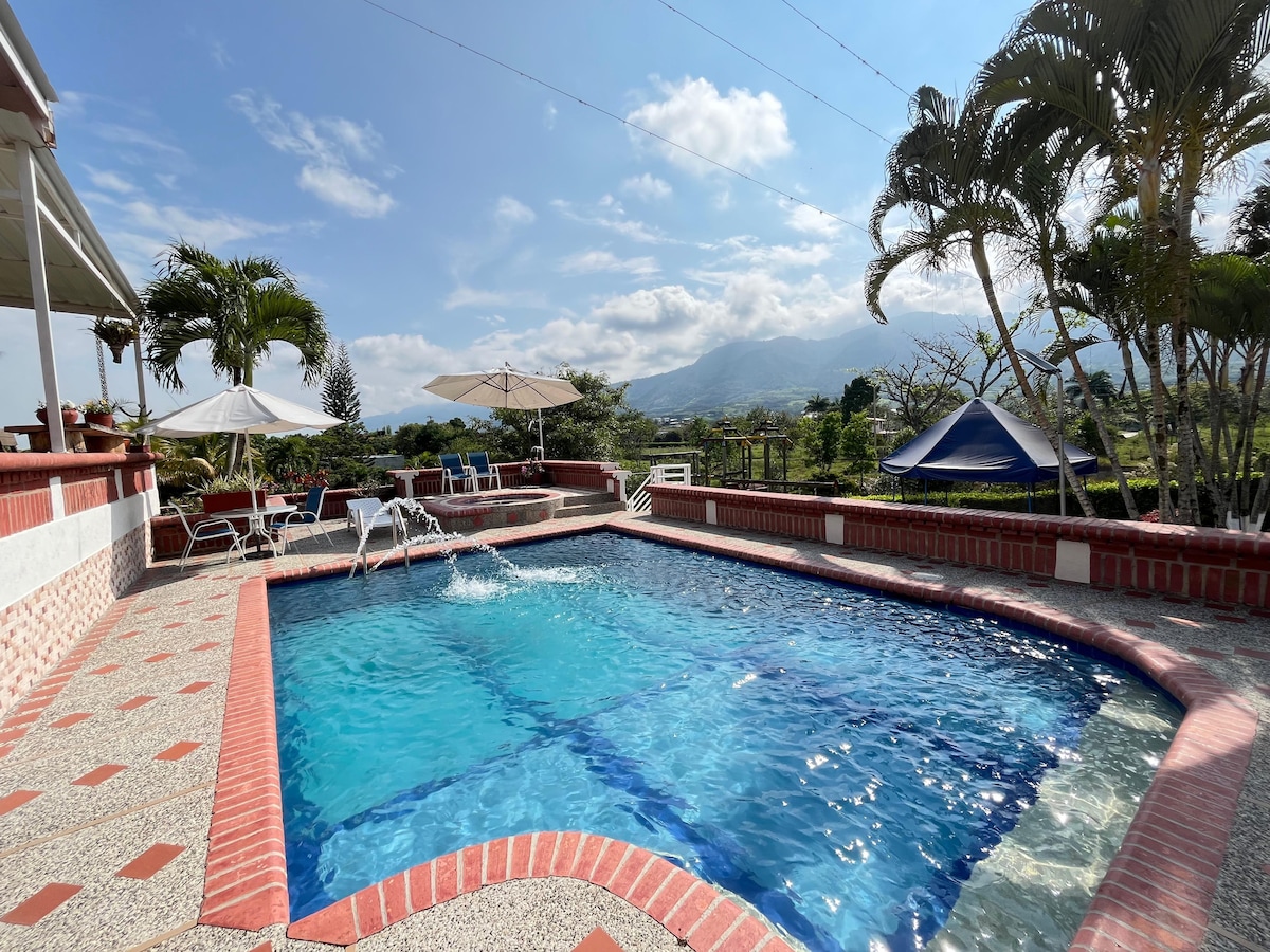 An inviting swimming pool is surrounded by a spacious patio. Sun loungers and umbrellas are positioned nearby, offering shade and comfort. Lush greenery and distant mountains create a serene backdrop under a partly cloudy sky.