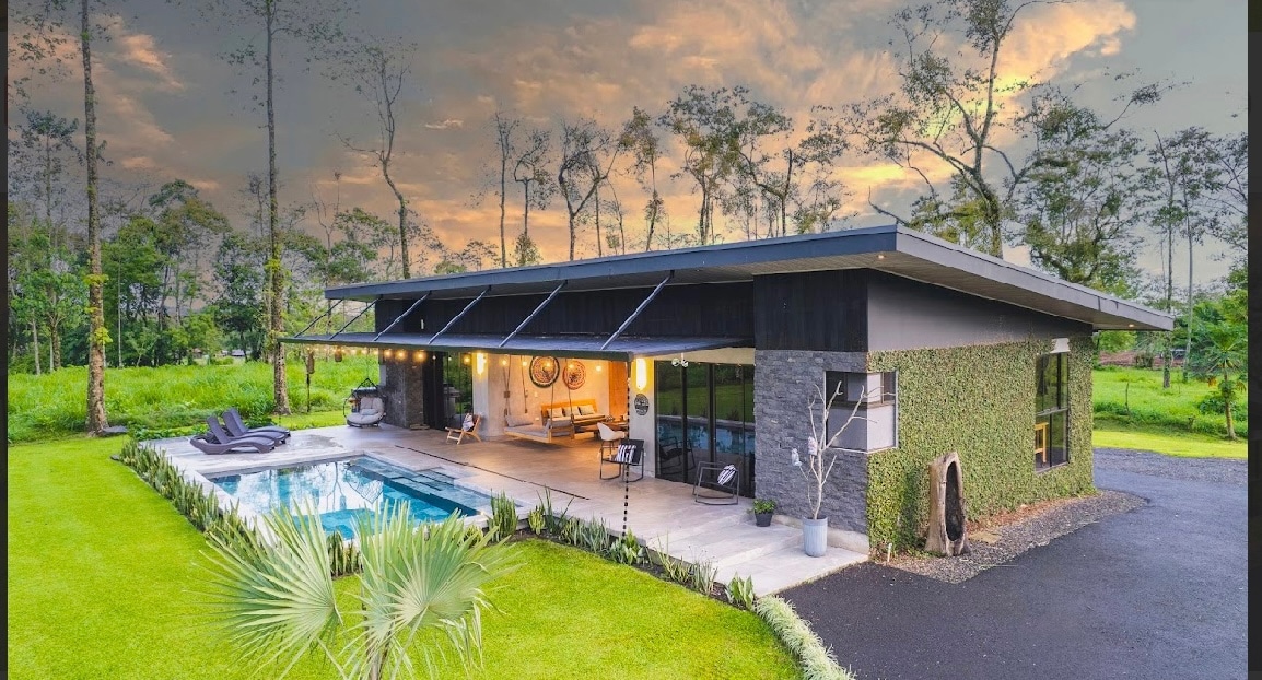 The contemporary vacation home is framed by lush greenery and a tranquil pool. Large windows and doors connect the indoor and outdoor living areas, while comfortable seating is arranged around the pool. The landscape showcases trees and open fields, enhancing the serene ambiance.