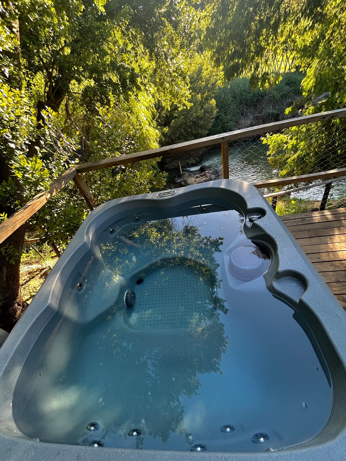 A hot tub is positioned on a wooden deck, surrounded by lush green foliage. Sunlight reflects on the water's surface, creating a serene atmosphere. In the background, a peaceful river can be seen, adding to the natural setting.