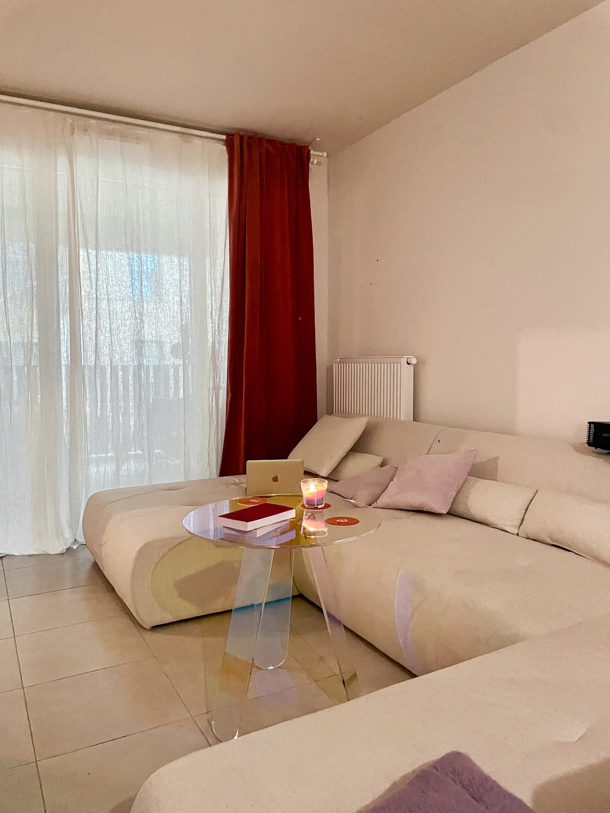 An inviting seating area features a neutral-colored sectional sofa adorned with soft cushions. A modern, transparent side table holds a small candle and a stack of books. Sheer curtains gently diffuse natural light from a nearby window, enhancing the relaxed ambiance.