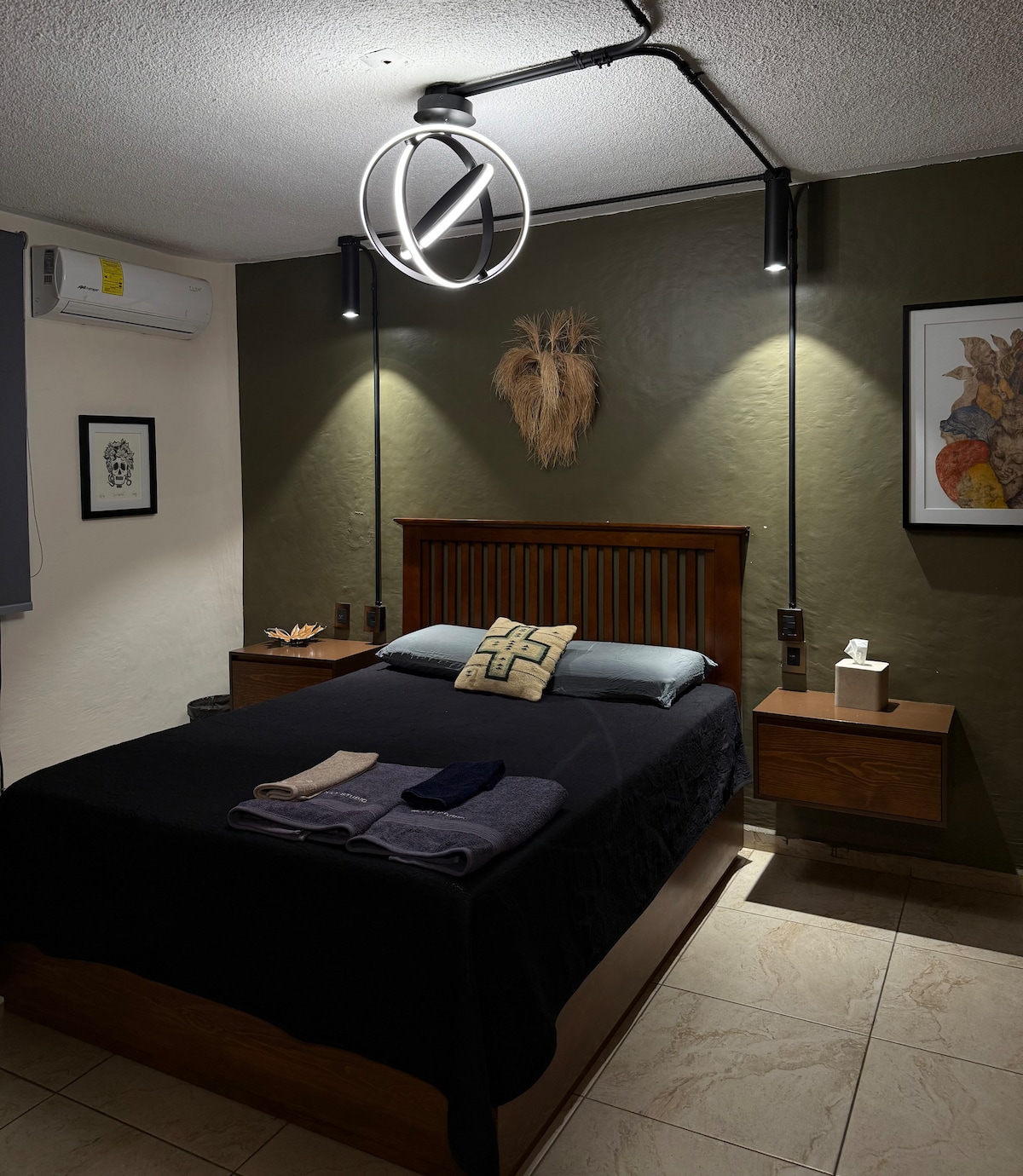 A cozy bedroom features a wooden bed with a dark blanket and decorative pillows. Wall-mounted lighting and minimalist decor create a calming ambiance. Nightstands on either side provide additional space, while the air conditioning unit is discreetly positioned above.