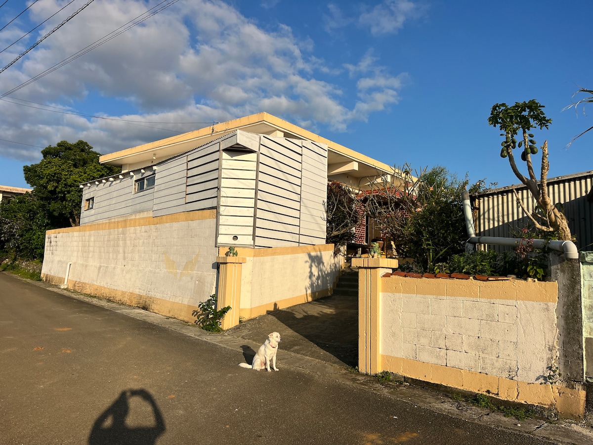A single-story house is depicted, featuring a light-colored wooden exterior and a welcoming entrance path. Lush greenery surrounds the property, while a pet dog sits calmly in the foreground, contributing to the peaceful scene. The clear blue sky enhances the inviting atmosphere.