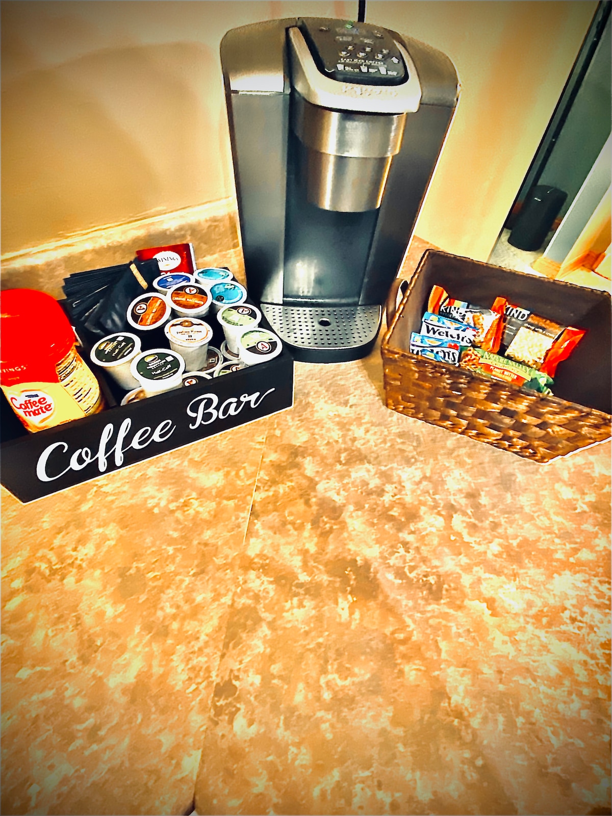 A coffee station is visible featuring a Keurig coffee maker alongside a selection of K-Cups organized in a black box labeled 'Coffee Bar.' A woven basket holds assorted snacks, providing convenience for guests.