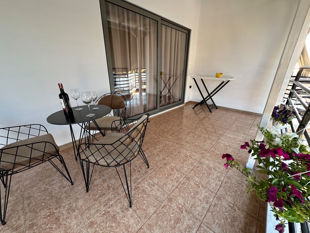 A balcony area features a stylish round table with two metallic chairs and a bottle of wine alongside glasses. A folding table is positioned nearby, while potted flowers add a touch of color, creating a pleasant outdoor setting.